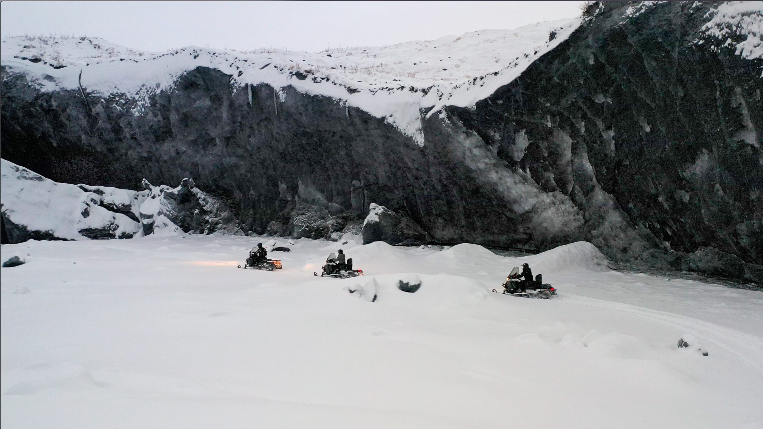 line of snowmobilers with headlights on near giant ice wall